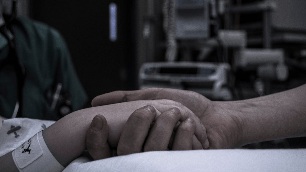 Man holding his dying wife's hand in a hospital bed while she is lying unconscious